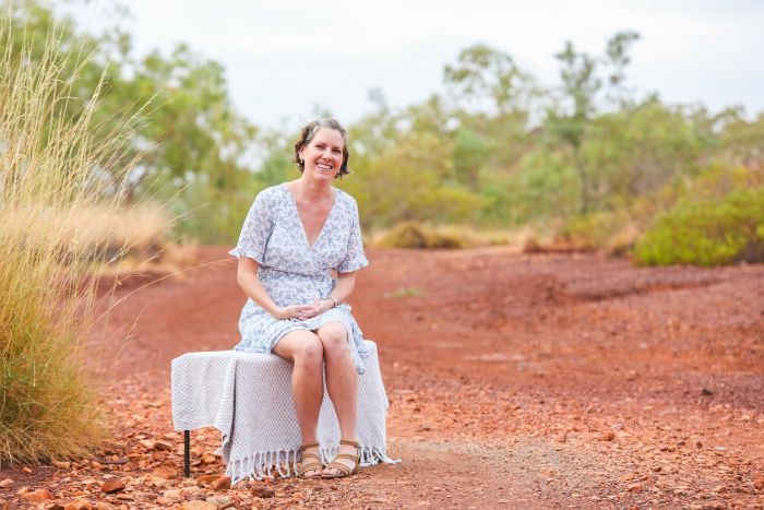 A woman in a floral blue and white dress sits on a bench in the Australian outback. 