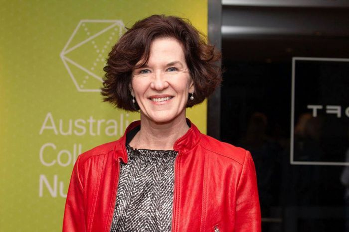 A middle aged woman with short brown hair stands in a red jacket in front of a green sign. 