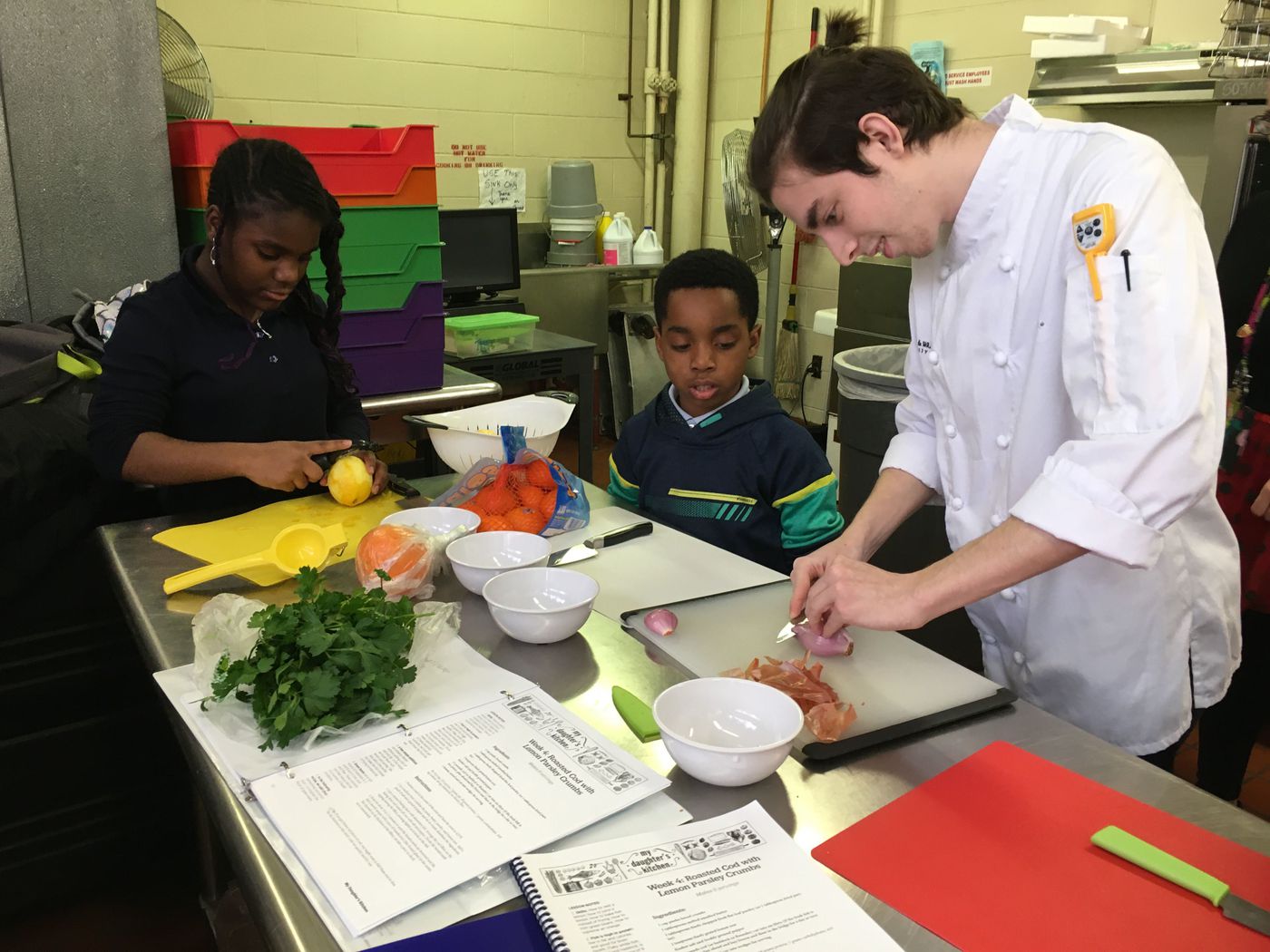 Miracle Brown, left, removes the zest from a lemon, and Shamar Ross watches as culinary student Aidan Dalton demonstrates how to mince a shallot, at the My Daughter's Kitchen after school cooking class at James Lowell Elementary School in North Philadelphia.