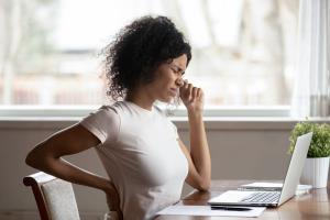 a woman standing in front of a laptop: Right now, many more people find themselves working from home. However, home offices have not necessarily been designed with ergonomics in mind, which can lead to back pain. Luckily there are easy ways to keep this at bay.